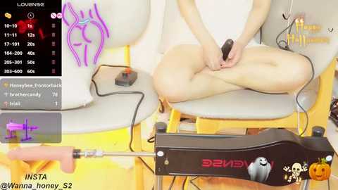 Media: A video depicts a nude woman with fair skin, sitting on a white chair, using a black vibrator on her genitals. The background features a yellow wall, and a smartphone screen displaying a low-energy warning.