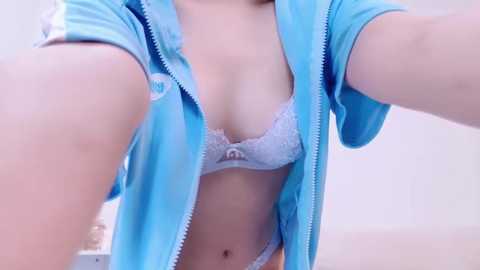 Media: Video of a light-skinned woman wearing a blue jacket open to reveal a white lace bra, capturing her midsection and upper torso in a selfie.