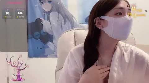 Media: Video of an Asian woman with long black hair, wearing a white surgical mask and robe, sitting in a sterile medical room with a poster of a woman with long hair.