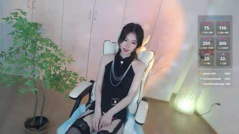 Media: A video of an Asian woman with pale skin and black hair, wearing a black sleeveless top and thigh-high stockings, sitting in a white gaming chair in a minimalist room.