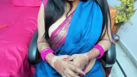 Media: Video of a woman with long black hair, wearing a blue saree with a pink blouse, intricate henna designs on her hands and arms. She sits on a black chair in a room with pink bedding and a yellow potted plant.