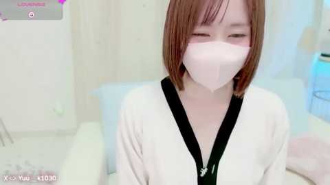 Media: Video of an East Asian woman with short brown hair and fair skin, wearing a white mask and black top, seated indoors with a blurred background.