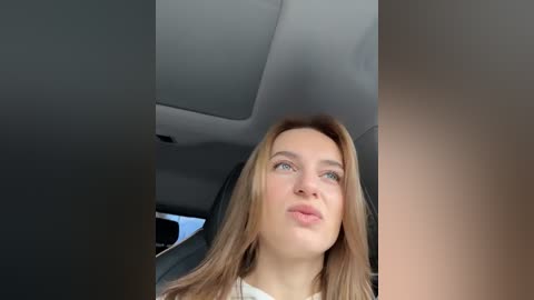 Media: Video of a young woman with long, straight, light brown hair, light skin, and blue eyes, seated in a car with gray interior, looking thoughtful.