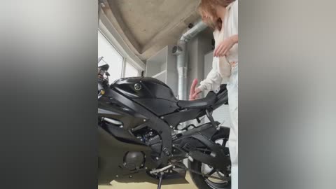 Media: Video of a woman in a white shirt and jeans examining a black sport motorcycle with fairings, inside a garage with concrete walls and industrial ceiling.