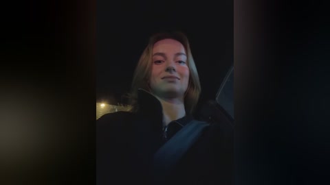 Media: Video of a woman with shoulder-length blonde hair, wearing a black coat, seen through a car window at night, dimly lit, with a blurred background.