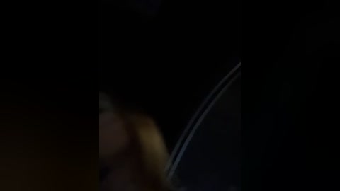 Media: This is a dimly lit video with a blurred figure, likely a woman with blonde hair, seen from behind, partially obscured by a dark background. The image suggests a nighttime setting, possibly in an urban environment.