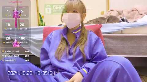 Media: Video of a female nurse with blonde pigtails, wearing a mask and purple scrubs, sitting on a hospital bed, surrounded by medical equipment and a green wall.