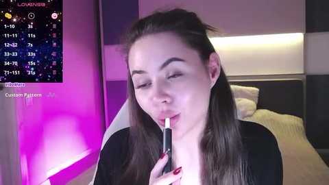 Media: Video of a young woman with long dark hair, wearing a black top, seductively licking a lollipop. Background shows a modern bedroom with purple and white lighting, a bed, and a TV screen displaying a calendar.