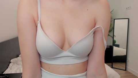 Media: A close-up video of a fair-skinned woman with medium-sized breasts wearing a light grey bra and matching panties. The background features a modern bedroom with a mirror and a bed.