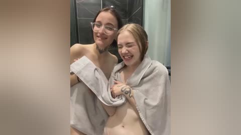 Media: Video of two young women, one with glasses and fair skin, the other with blonde hair, smiling, wrapped in towels in a tiled shower.