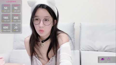 Media: Video of an East Asian woman with long black hair, wearing white bunny ears, glasses, a white off-shoulder top, and a black choker. She sits in a white room with a gray monitor displaying stats in the background.