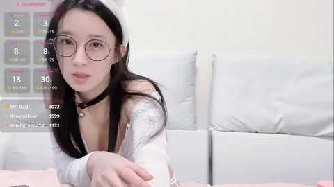 Media: A video of an East Asian woman with long black hair, wearing glasses, a white headband, and a choker, lying on a white bed, with a digital overlay showing her stats.