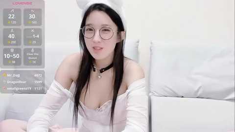 Media: A video of an Asian woman with long black hair, wearing a white bunny hat, off-shoulder white dress, and glasses, lying on a bed, with a digital health stats overlay.