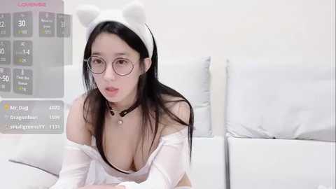 Media: Video of a young Asian woman with long black hair, wearing a white off-shoulder dress and cat ear headband, sitting on a white couch.