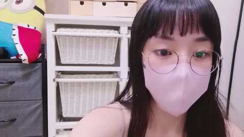 Media: Video of an Asian woman with long black hair, wearing round glasses and a white mask, standing in front of white shelves with baskets and colorful folders in a bright room.