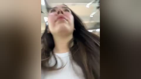 Media: A blurry video of a woman with long, dark hair and a white shirt, taken from a low angle, looking upward. The background is indistinct, suggesting an indoor setting.