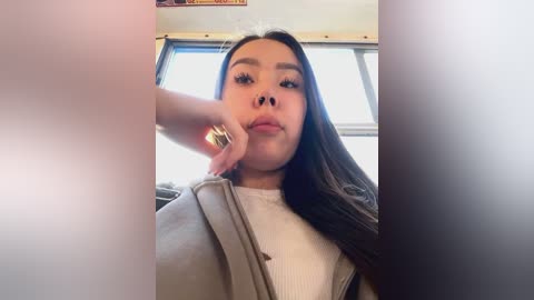 Media: Video of a young Asian woman with long black hair, wearing a beige jacket over a white top, seated in a car with sunlight streaming in through the window.