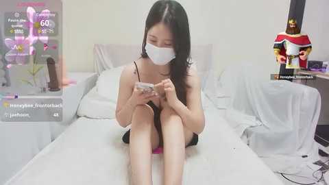 Media: A video of an Asian woman with long black hair, wearing a face mask and black camisole, sitting on a white bed, playing a smartphone game.