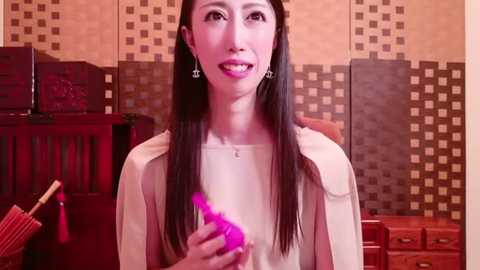 Media: A video of a smiling East Asian woman with long black hair, wearing a cream blouse, holding a pink vibrator in a home setting with wooden furniture and tiled walls.