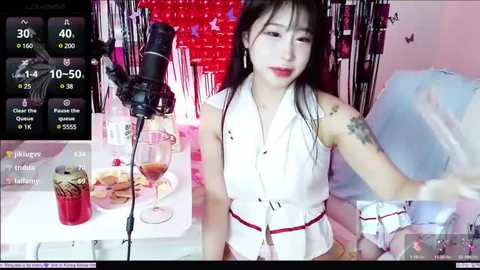 Media: Video of a young Asian woman with long black hair, wearing a sleeveless white blouse and red skirt, smiling while recording herself with a camera on a white table with wine glasses and food.