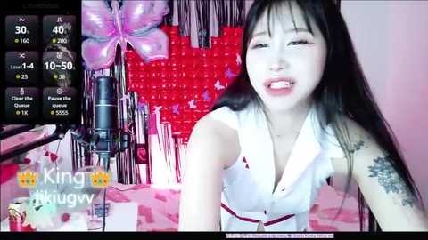 Media: A video of a young Asian woman with long black hair, wearing a sleeveless white top, smiling while holding a microphone in a room decorated with red hearts and pink tinsel.