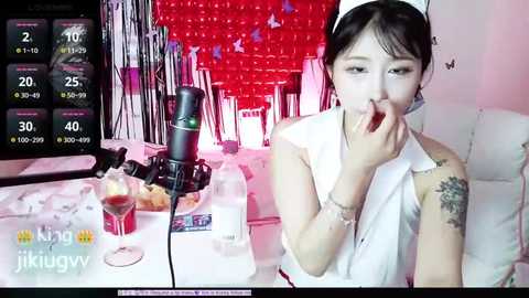 Media: Video of an Asian woman with black hair, wearing a white sleeveless top, eating a piece of food while sitting at a table with wine glasses, a microphone, and a monitor displaying a countdown.