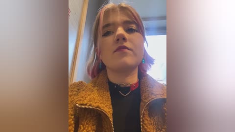 Media: Video of a young woman with short, dyed pink and blonde hair, wearing a textured mustard jacket over a black top, and a heart-shaped necklace, indoors.