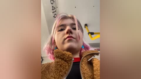 Media: Video of a young woman with pink-dyed hair, light skin, and a tan, fuzzy jacket. She wears blue earrings and a black shirt. Background includes a yellow handrail and white wall with black text.