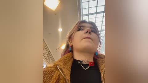 Media: Video of a young woman with pale skin, light pink hair, and blue eyes, wearing a brown fuzzy jacket, black top, and colorful earrings. She gazes upward, surrounded by modern, bright, and minimalistic architectural features.