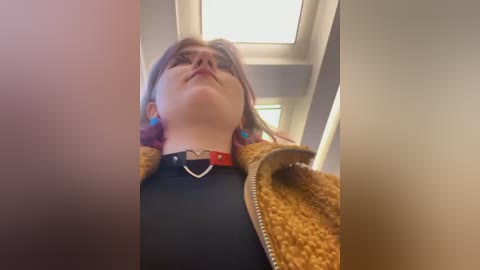 Media: A video of a young woman with glasses, pale skin, and light brown hair, wearing a heart pendant necklace and a mustard jacket, taken from a low angle, capturing a candid, slightly distorted view.