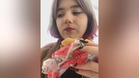 Media: A video of a young woman with shoulder-length, pastel pink hair, wearing a brown cardigan, eating a sandwich. Her expression is neutral, and the background is blurred.