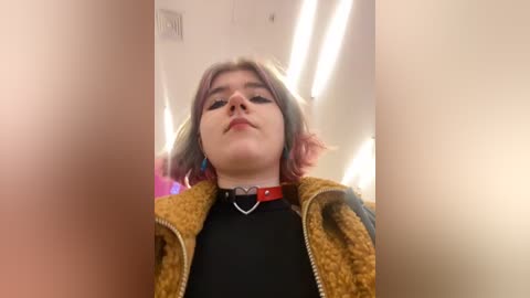 Media: Video of a young woman with light skin, shoulder-length wavy hair, wearing a black shirt, mustard-yellow fleece jacket, and a red collar with a heart pendant. She has prominent eye makeup and is indoors with fluorescent lights overhead.