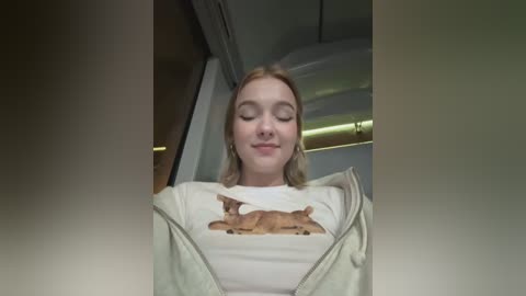 Media: A video of a young woman with fair skin, blonde hair, and closed eyes, wearing a white t-shirt with a brown bear graphic and a beige jacket. She's seated in a vehicle's interior, with a beige ceiling and a window visible.