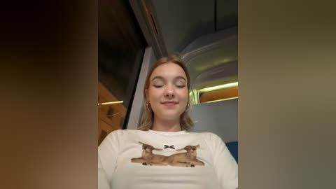 Media: Video of a young woman with fair skin, closed eyes, and a slight smile, wearing a white t-shirt with a reindeer graphic, seated in a dimly lit airplane cabin.