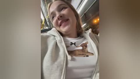 Media: Video of a young Caucasian woman with fair skin, smiling, wearing a white shirt with a deer graphic and a beige hoodie. Background shows a blurry interior with a yellow safety vest.