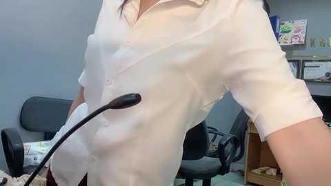 Media: Video of a woman in a white blouse, slightly unbuttoned, revealing cleavage. She has a microphone in her hand, standing in an office with gray walls, a desk, and a chair.