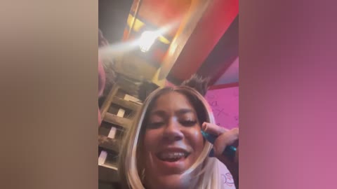 Media: Video of a joyful woman with light skin and shoulder-length blonde hair, wearing a light-colored shirt, holding her head in a playful manner. Background features a dark room with a cat tower and pink lighting.