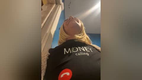 Media: A video of a woman with blonde hair, wearing a black \"Money calling\" shirt, standing in a room with blue walls and a ceiling light.