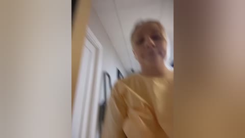 Media: A blurry video of a middle-aged woman with short blonde hair, wearing a yellow top, standing in a hallway with white walls and closed doors. The image is slightly out of focus.