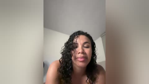 Media: A video of a light-skinned woman with curly black hair, eyes closed, and a serene expression, standing in a minimalist room with white walls and a ceiling.