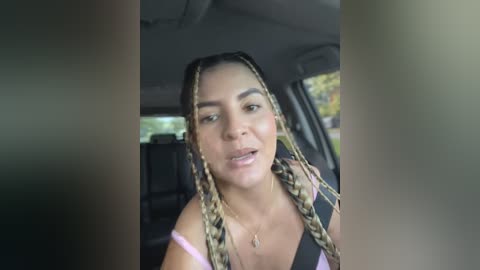 Media: Video of a young Black woman with long braided hair, light skin, wearing a black and pink top, sitting in a car.