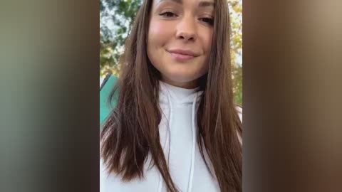 Media: A video of a smiling young woman with long brown hair, wearing a white turtleneck, standing outdoors with blurred greenery in the background.