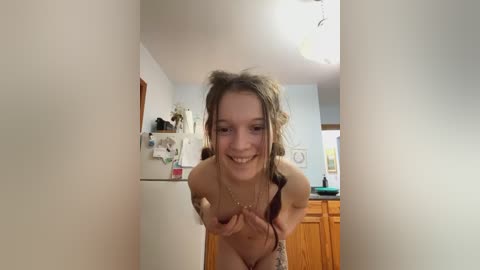 Media: Video of a smiling, nude young woman with light skin and long, curly brown hair. She is bent over slightly, hands on her breasts, in a cozy kitchen with wooden cabinets and a fridge in the background.