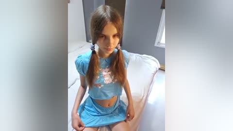 Media: Video of a young Caucasian girl with long, straight light brown hair in pigtails, wearing a blue crop top and matching skirt, sitting on a bed in a gray room.