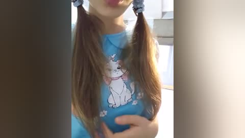 Media: Video of a young woman with long brown hair in two pigtails, wearing a blue t-shirt with a white cat graphic, holding her belly. Background shows a bright room with a white door.