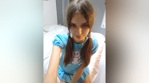 Media: Video of a young woman with light brown hair in pigtails, wearing a blue t-shirt with a cat graphic, seated on a bed in a minimalist bedroom.