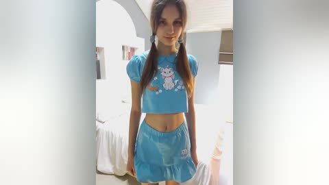 Media: Video of a young woman with fair skin, wearing a blue crop top and matching skirt adorned with a cartoon cat, posing indoors in a well-lit room.