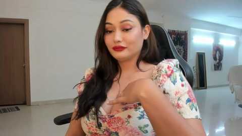 Media: Video of a woman with long black hair, wearing a floral dress, sitting on a black office chair in a modern, minimalist room with white walls and a large window.