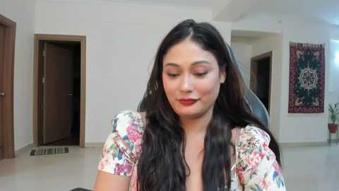 Media: Video of a young woman with long dark hair, wearing a floral dress, seated indoors, with white walls, a red tapestry, and closed doors in the background.