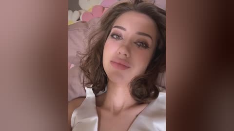 Media: Video of a young woman with wavy, shoulder-length brown hair, light skin, and full lips, wearing a white satin robe, lying on a pink bed with stuffed animals.